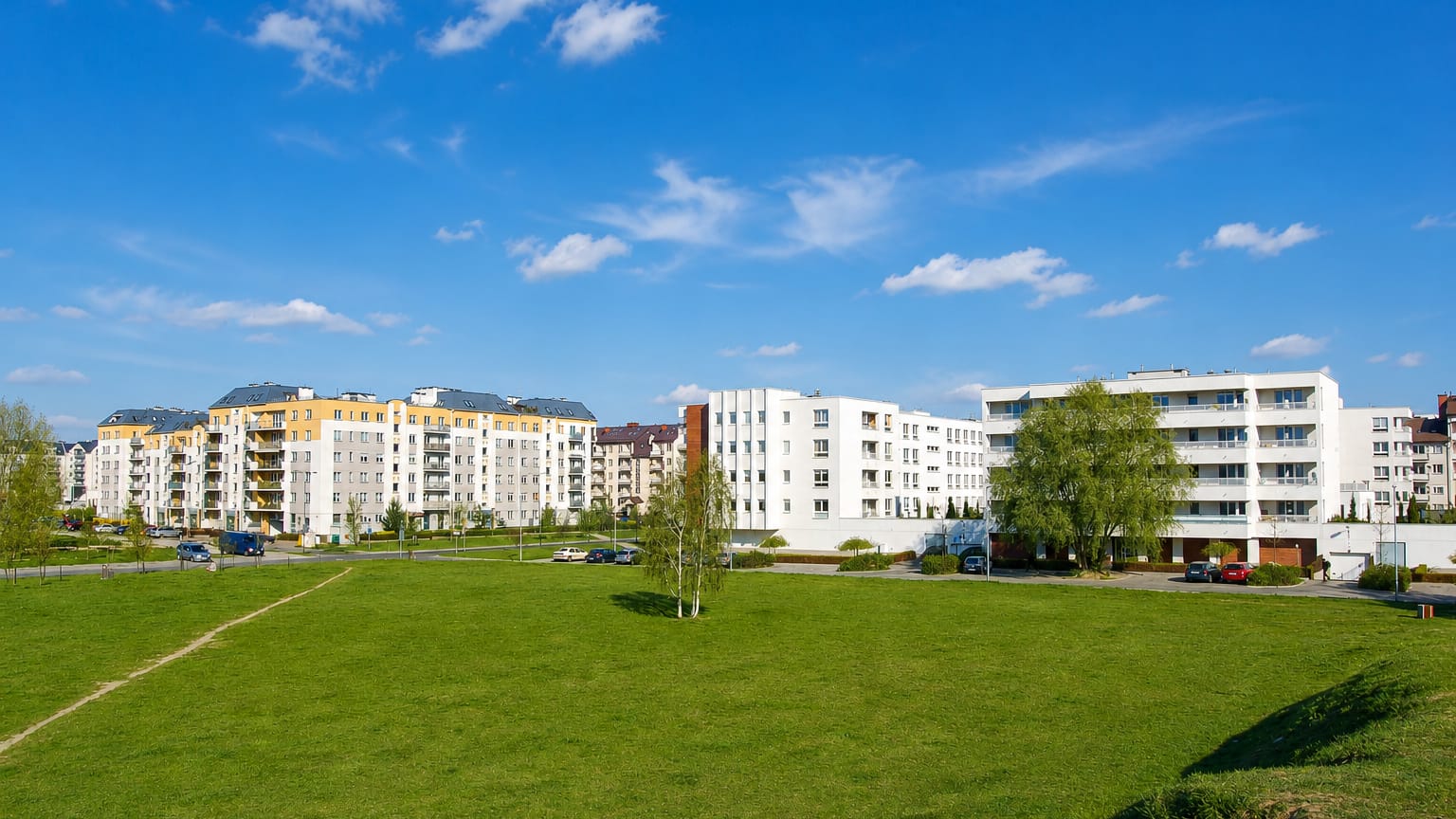Modern white and yellow apartment blocks behind a green park in Warsaw's Kabaty district under a blue sky.