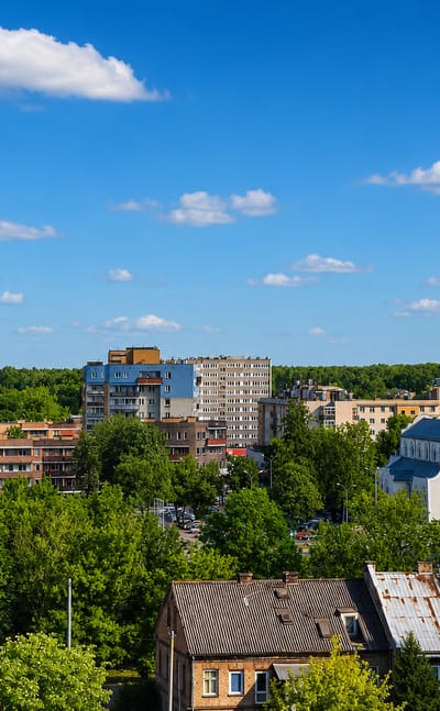 Bialystok skyline with a white church spire, apartment towers and an old timber house among trees.