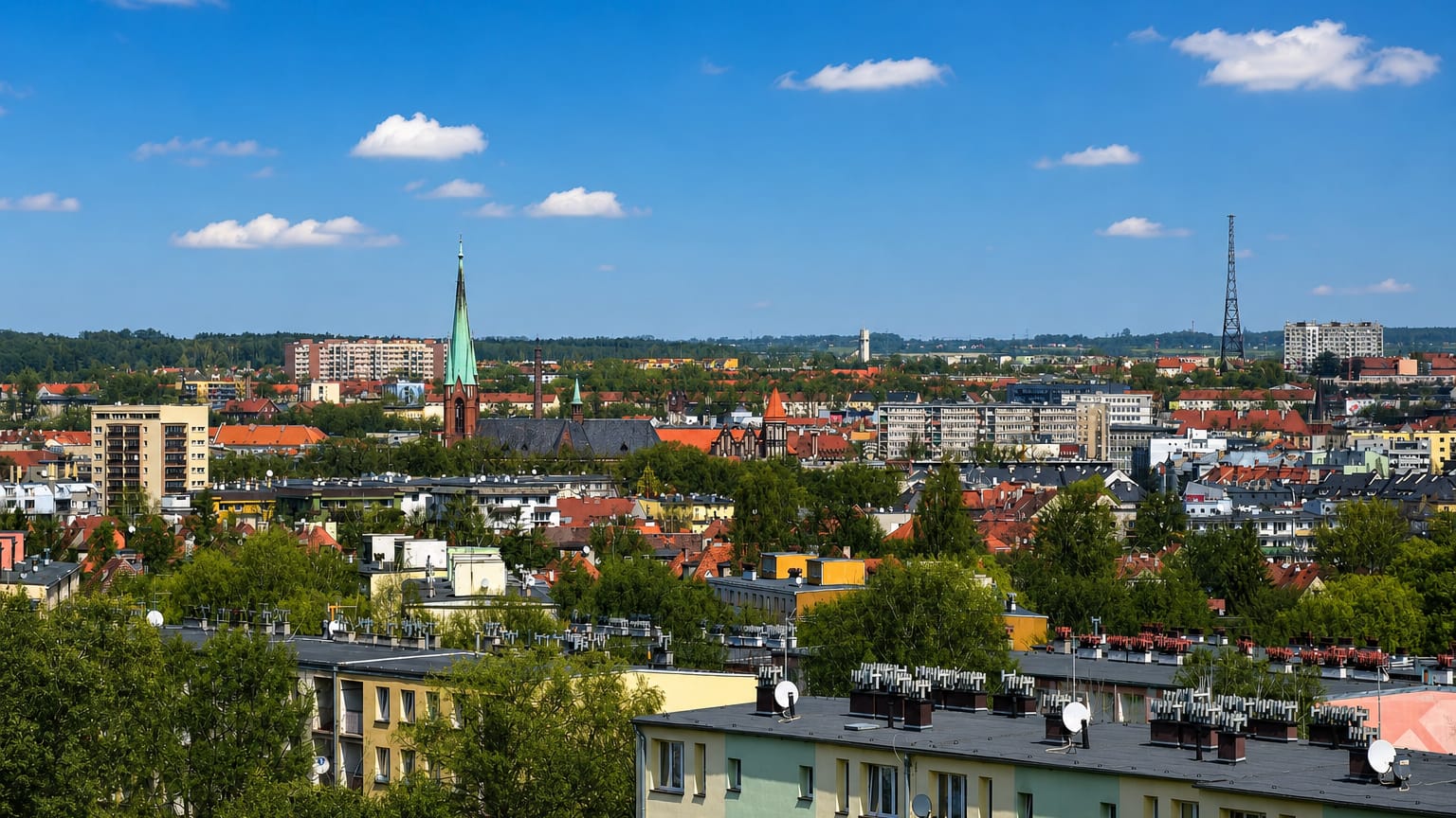 Gliwice townscape with a green church spire and a lattice radio tower rising above rooftops under a blue sky.