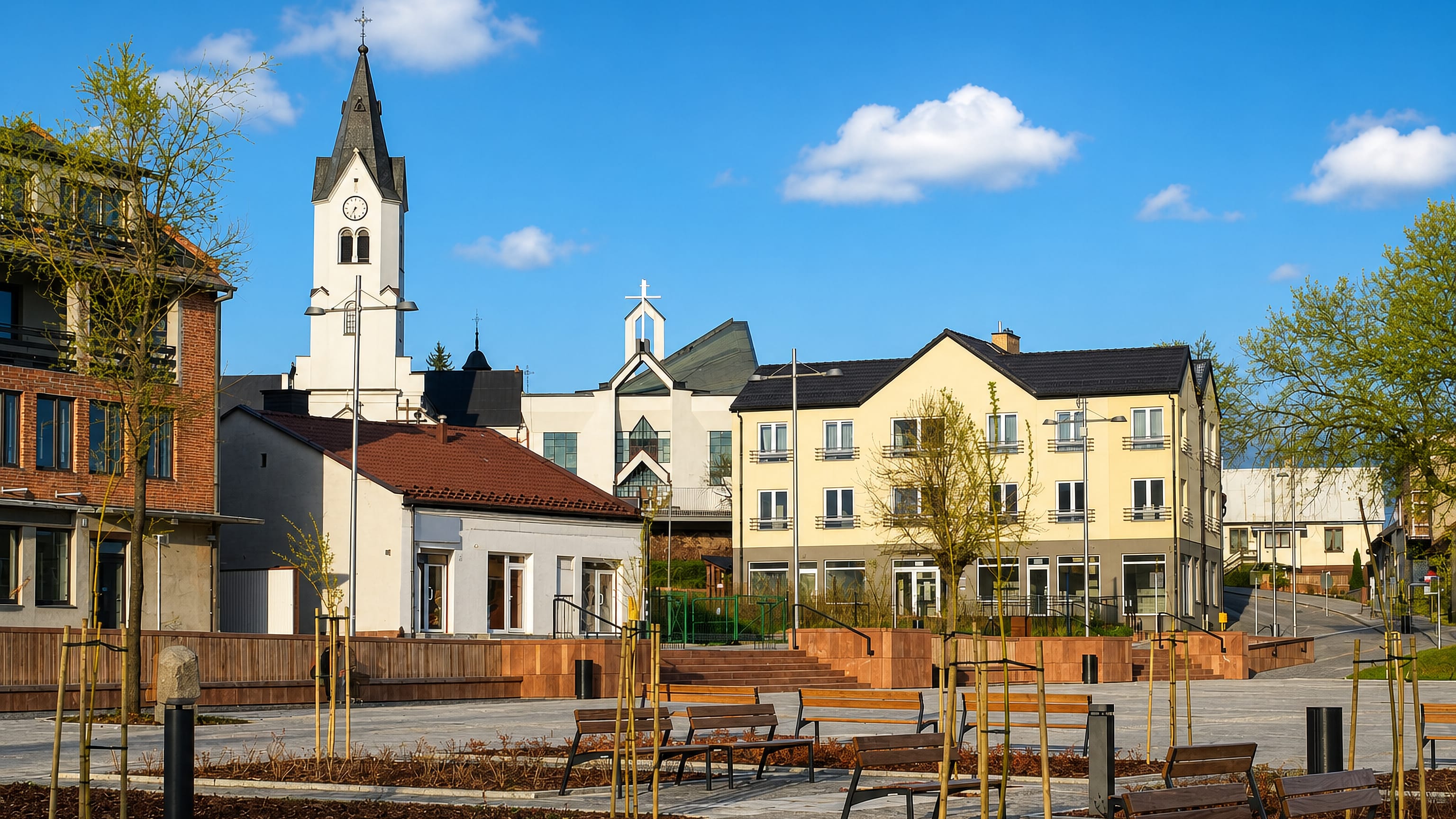 Renovated town square with benches and young trees below a white clock-tower church in Starachowice, Poland.