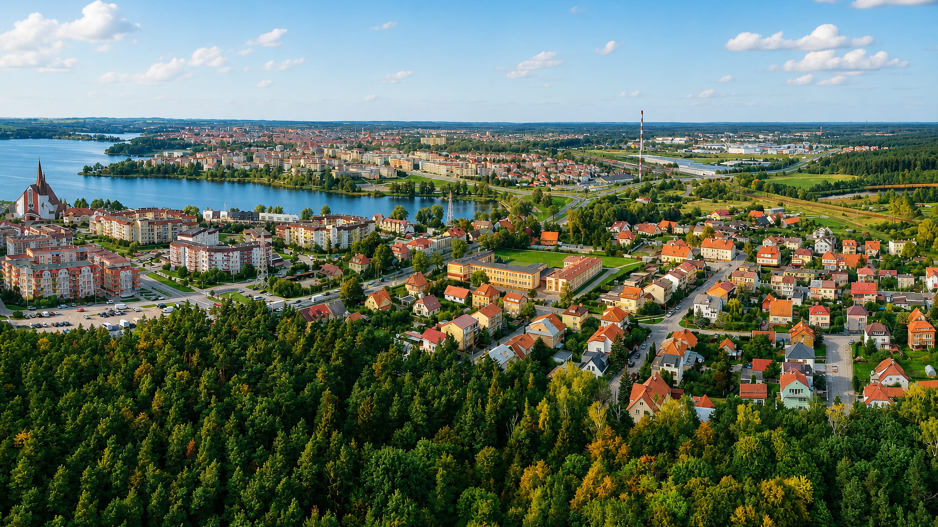 Aerial view over Elk's Szyba district, with forest and houses and the town's lake and brick church beyond.
