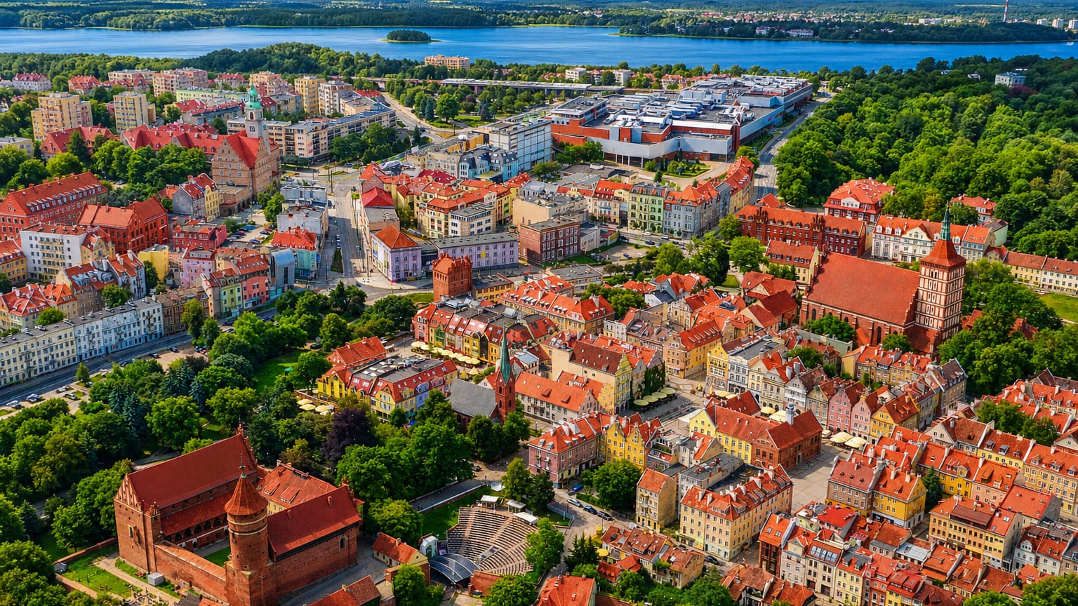Aerial view of Olsztyn's Old Town, its red-brick castle and Gothic cathedral, with a Masurian lake beyond.