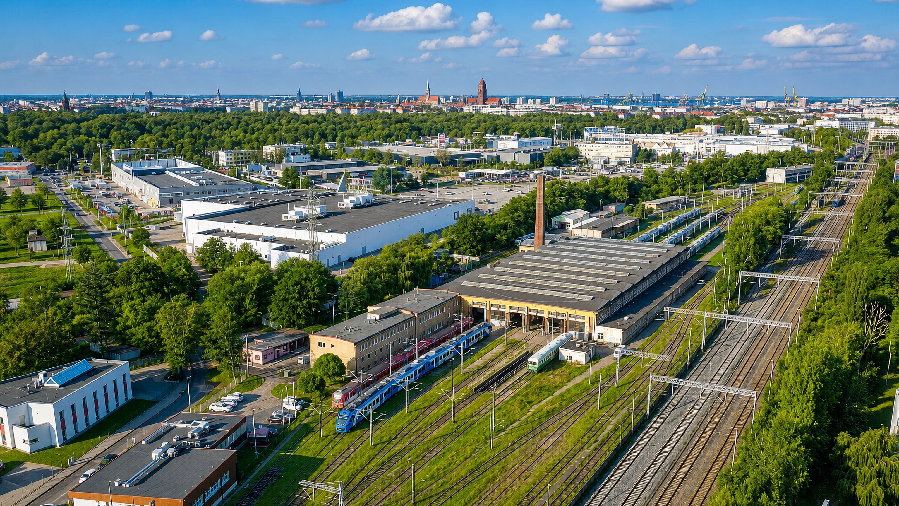 Aerial view of Pomorzany, Szczecin: rail depot sheds and sidings with the city skyline and port cranes beyond.