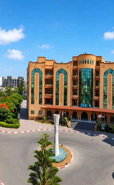 Ornate mid-rise building beside a twin-minaret mosque and a monument roundabout with palm trees in Gaza City.