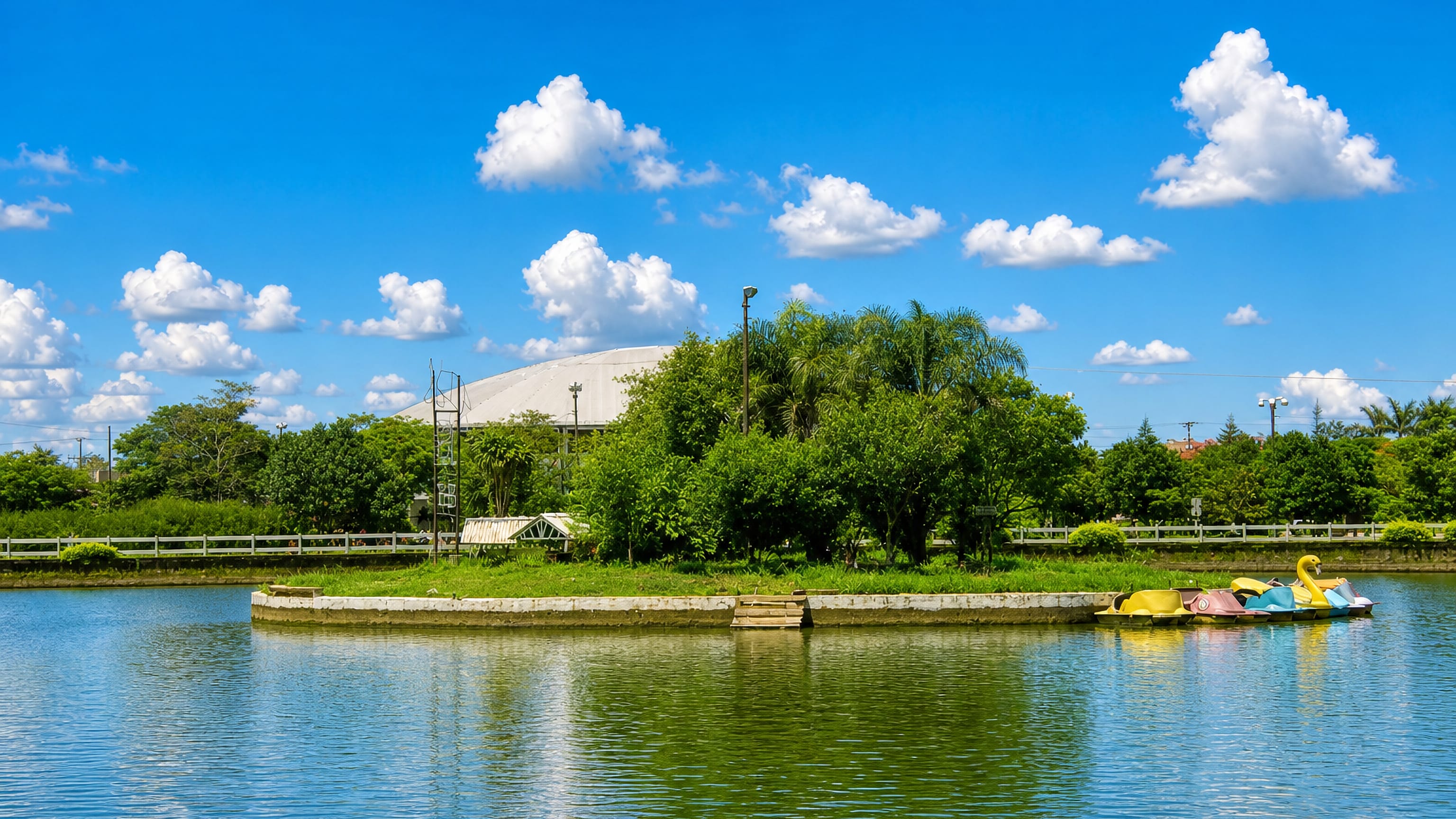 Pedal boats on a park lake in Pedro Juan Caballero, Paraguay, with an arena roof behind the trees