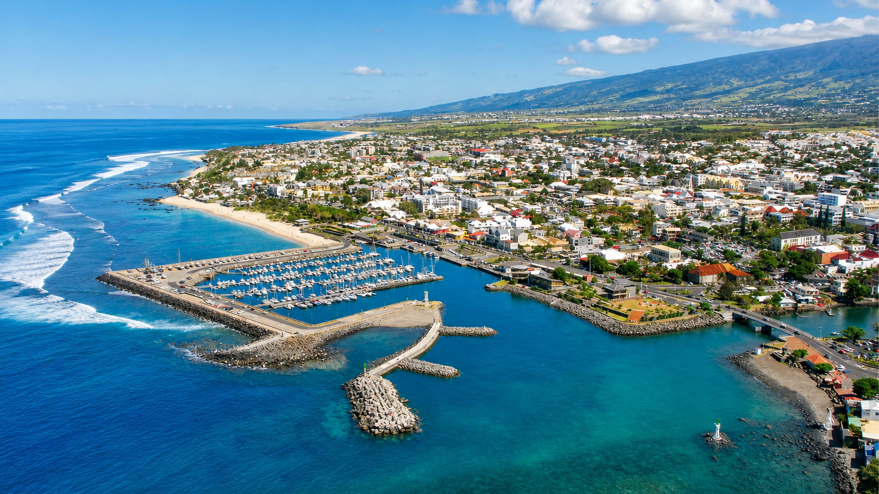 Aerial view of Saint-Pierre, Réunion, its breakwater-sheltered marina, beach and the island's mountains behind.