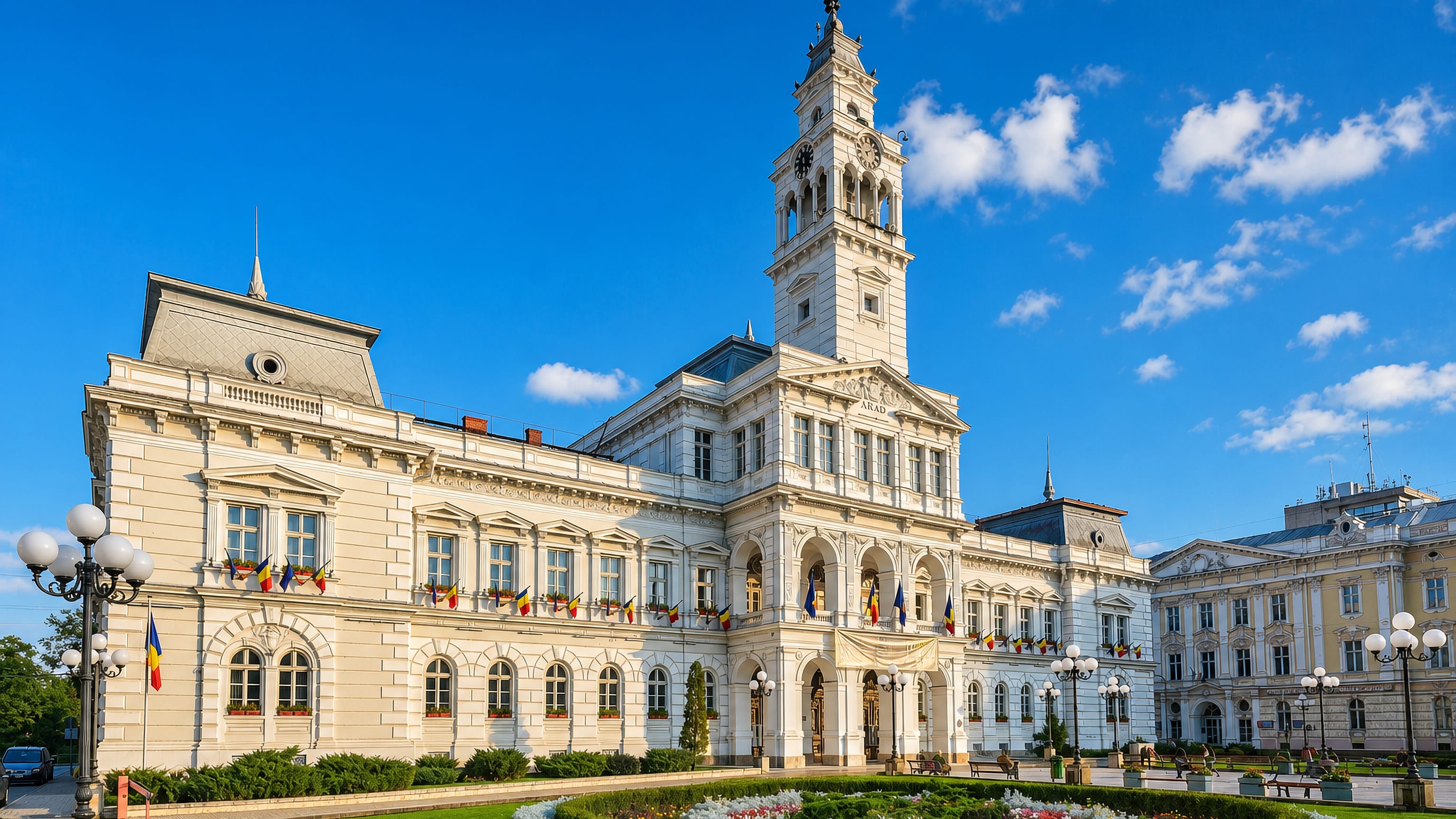 Arad City Hall in Romania, its clock tower and flag-lined neoclassical facade above a flowerbed-ringed square.