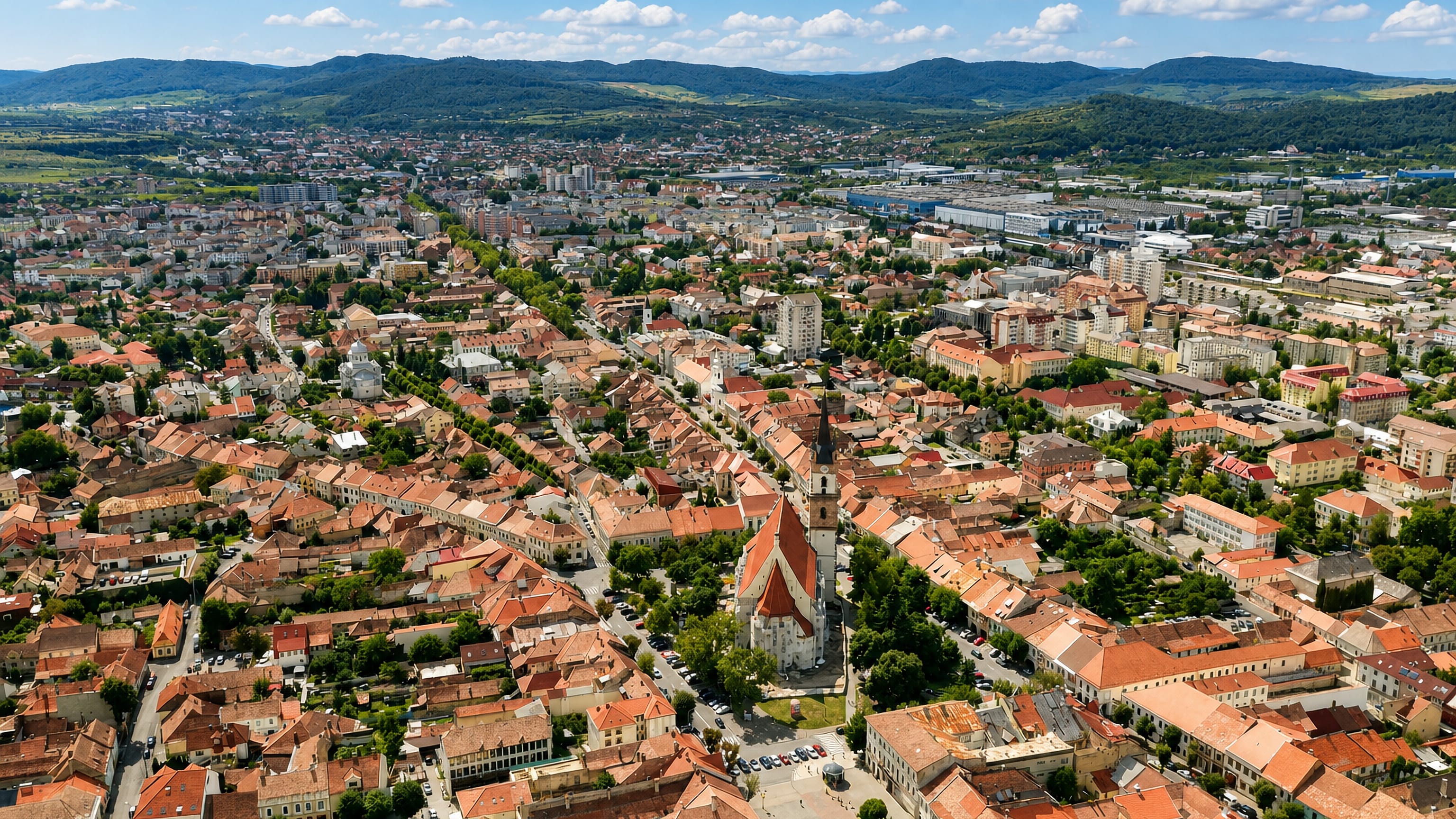 Aerial view of Bistrita, Romania, centred on the Saxon Evangelical church tower amid red-tiled roofs and hills.