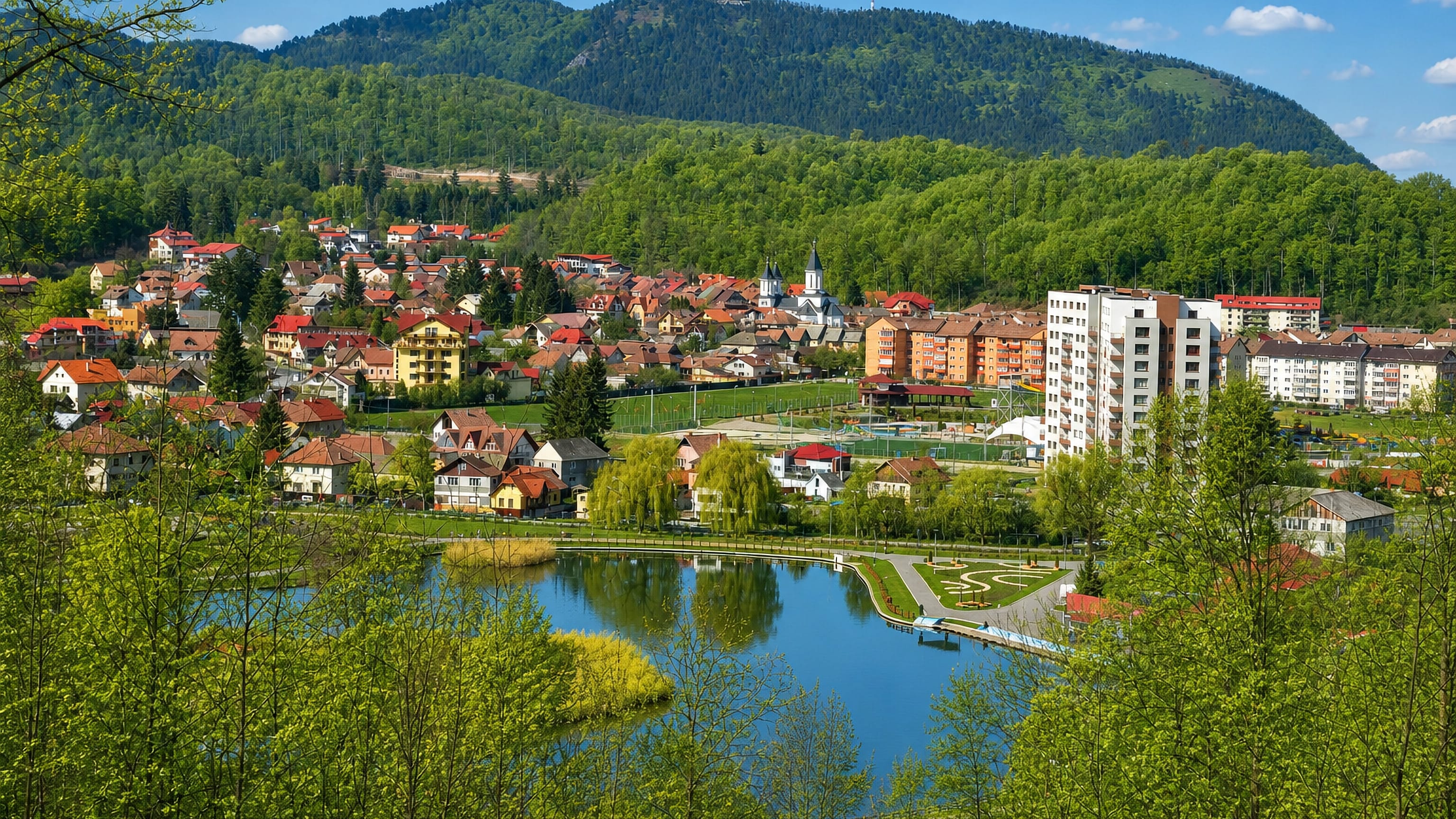 Houses and apartment blocks around the lake at Noua, Brasov, below a forested mountain topped by a mast.