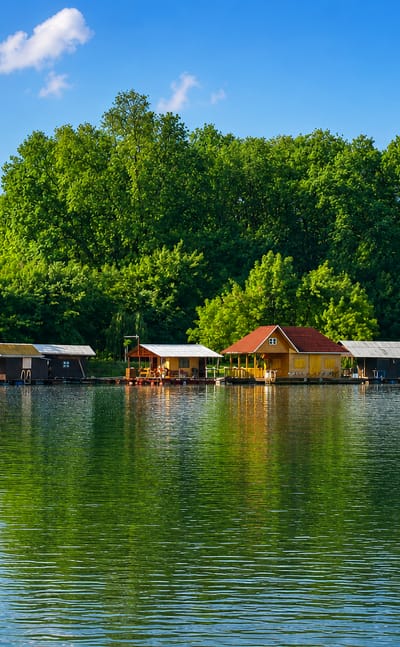 Wooden weekend cabins line the wooded, calm-water shore of Ada Medica river island in Belgrade under a blue sky.