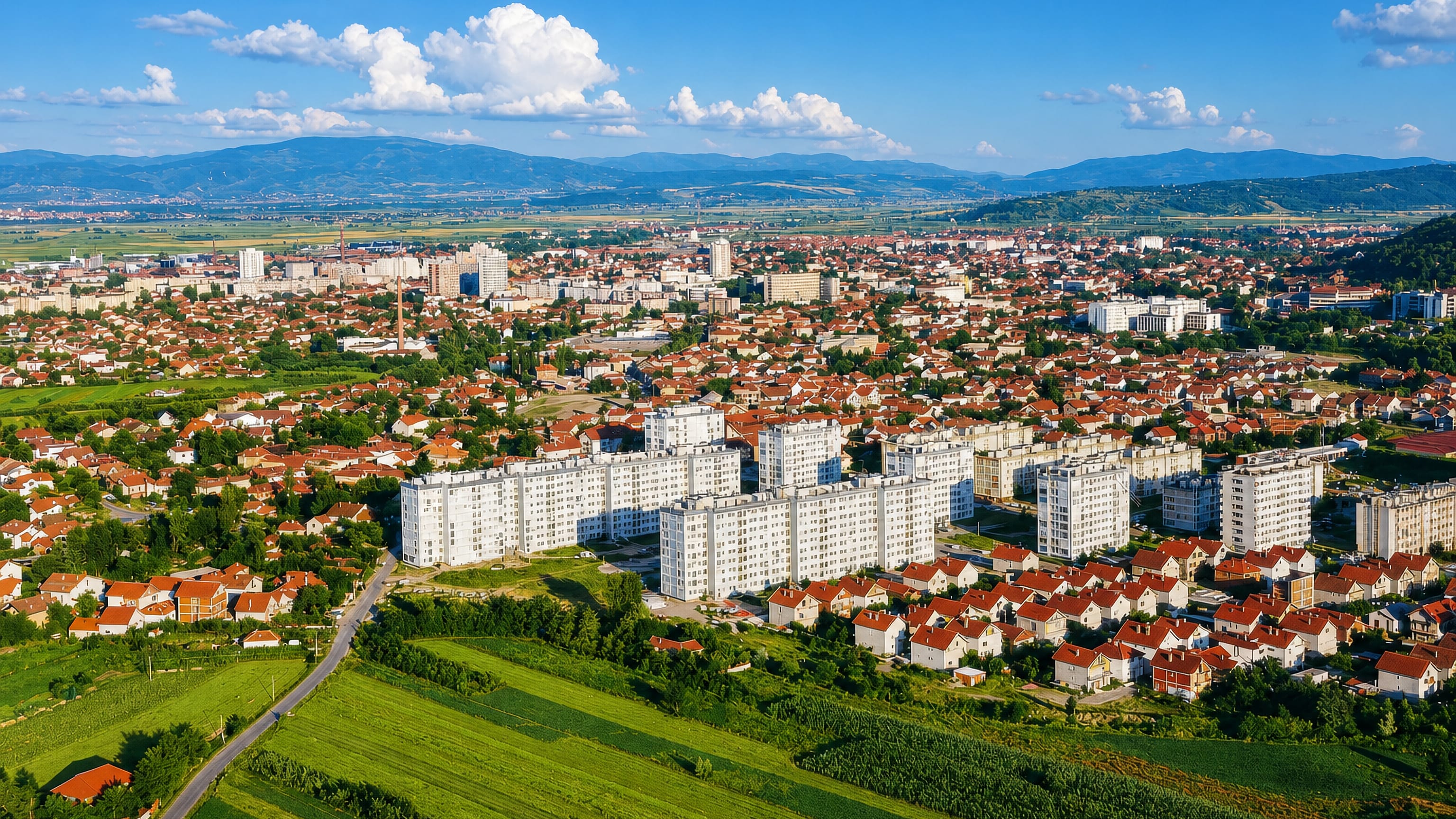 Aerial view of Leskovac, Serbia, with red-roofed houses, white apartment blocks and farmland below distant hills.