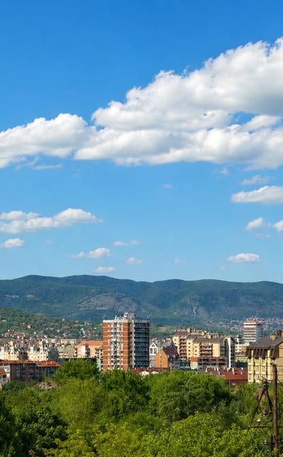 Apartment blocks and red-tiled houses of Niš, Serbia, spread below forested hills and distant mountains.