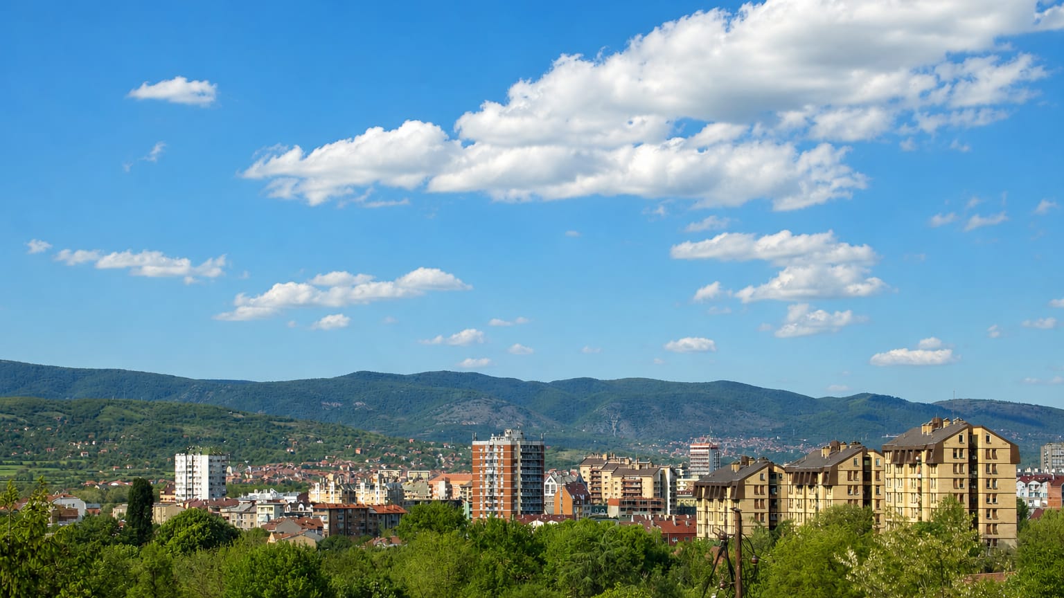 Apartment blocks and red-tiled houses of Niš, Serbia, spread below forested hills and distant mountains.