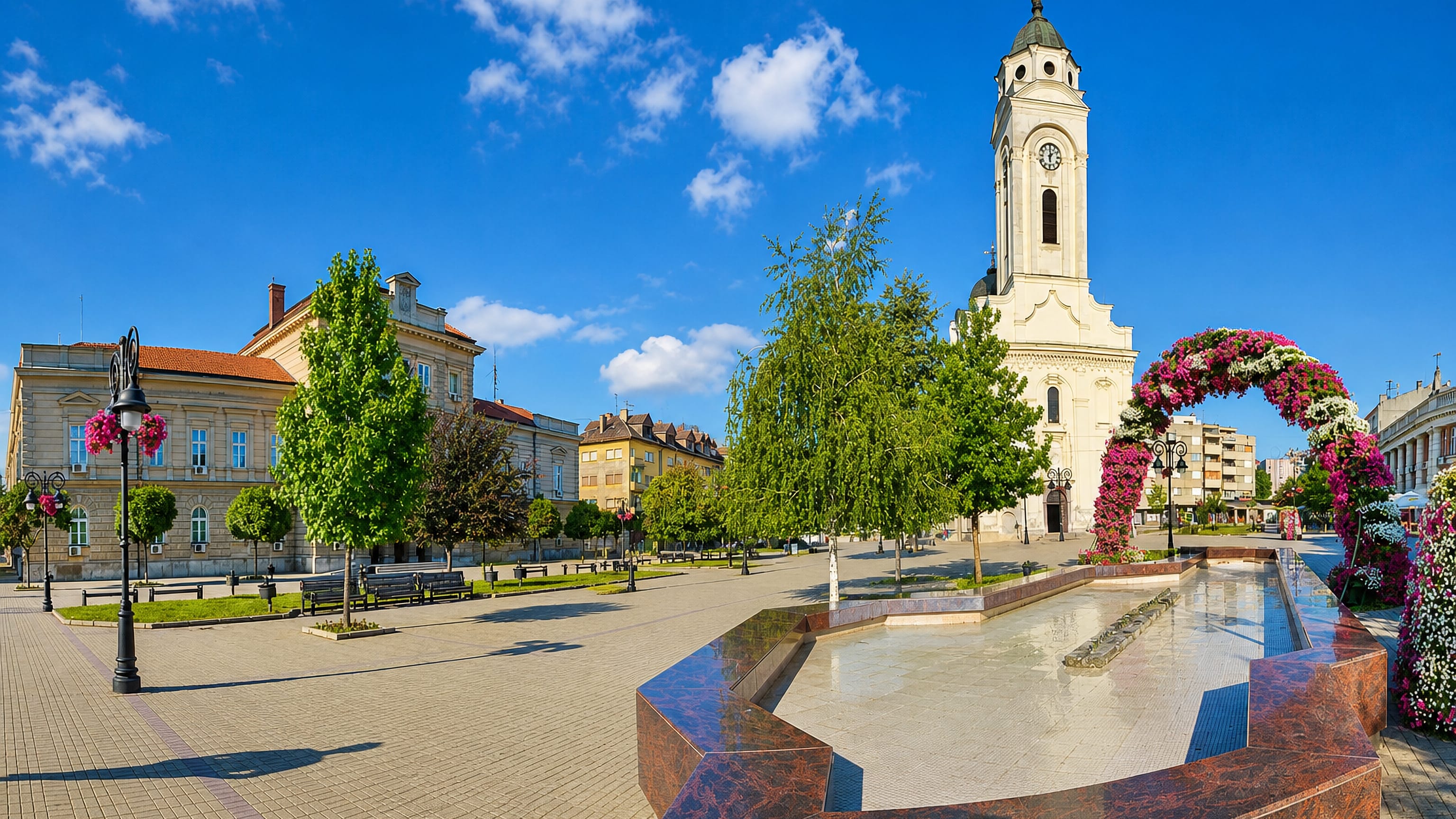 Smederevo's main square in Serbia, with a white clock-tower church, flower arch and stone fountain under blue sky