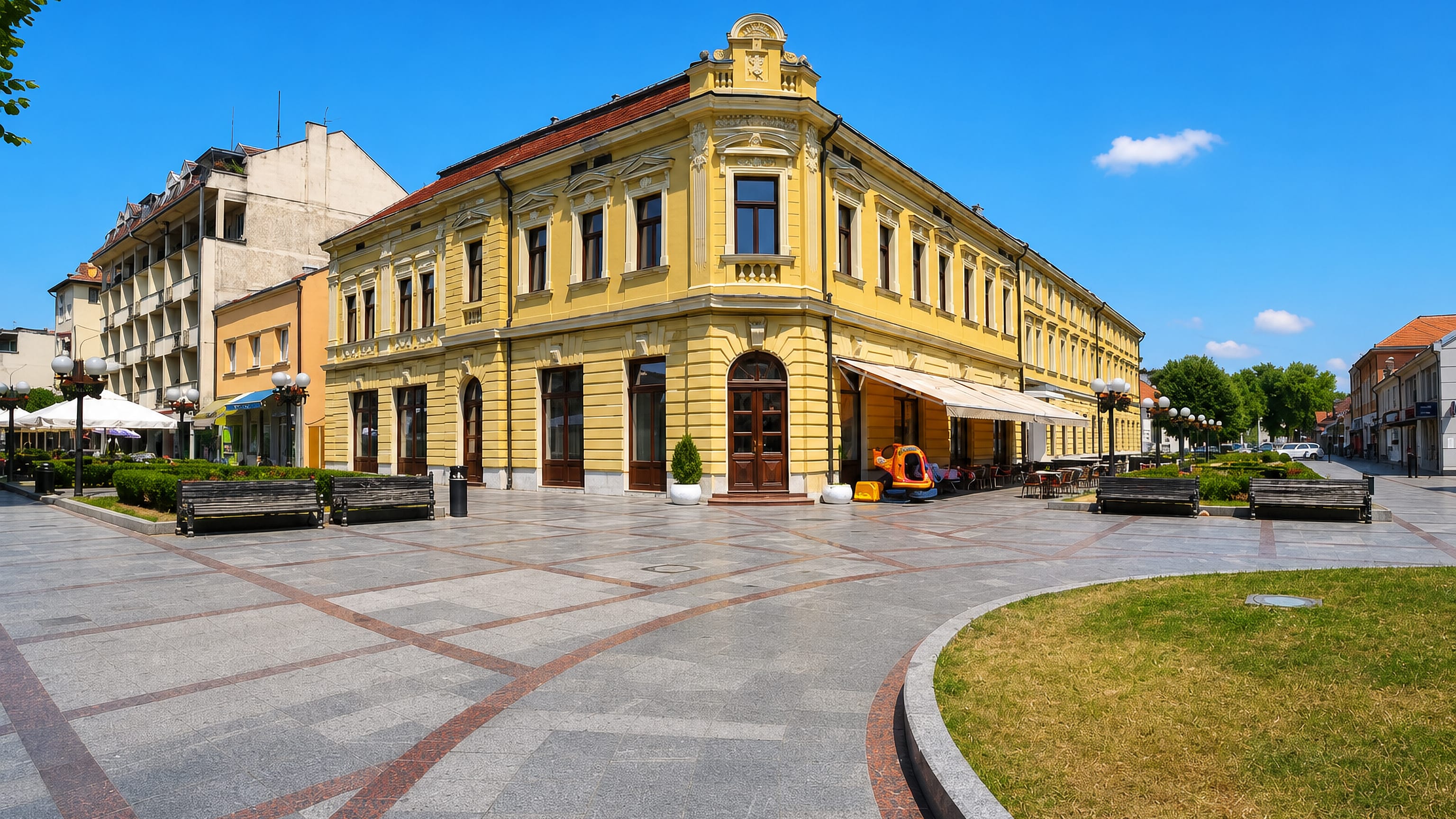 Valjevo's paved pedestrian square with a yellow historicist corner building and café terraces.