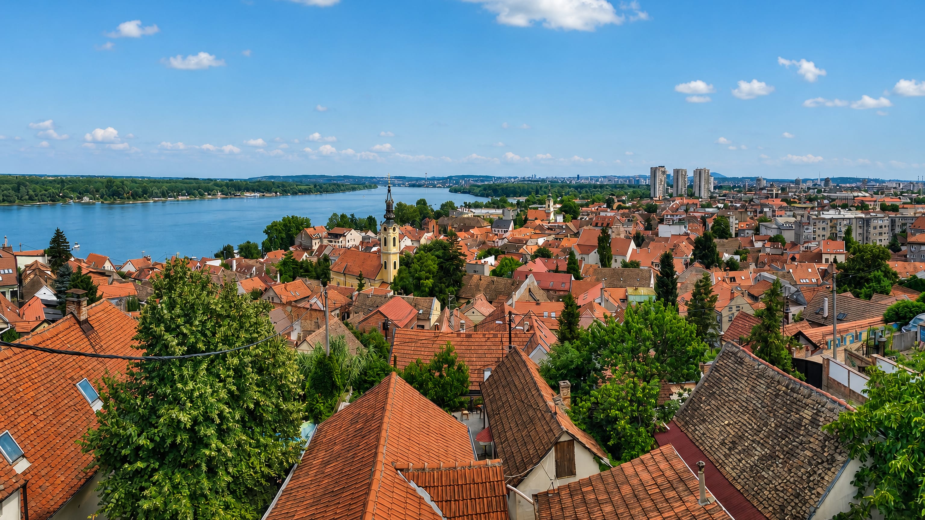 Red-tiled rooftops of Zemun sloping to the Danube, with a church tower and New Belgrade towers beyond.