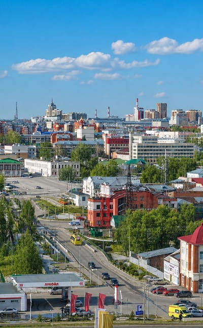 Wide daytime panorama of Barnaul, Russia, with historic rooftops and apartment towers beyond leafy streets.