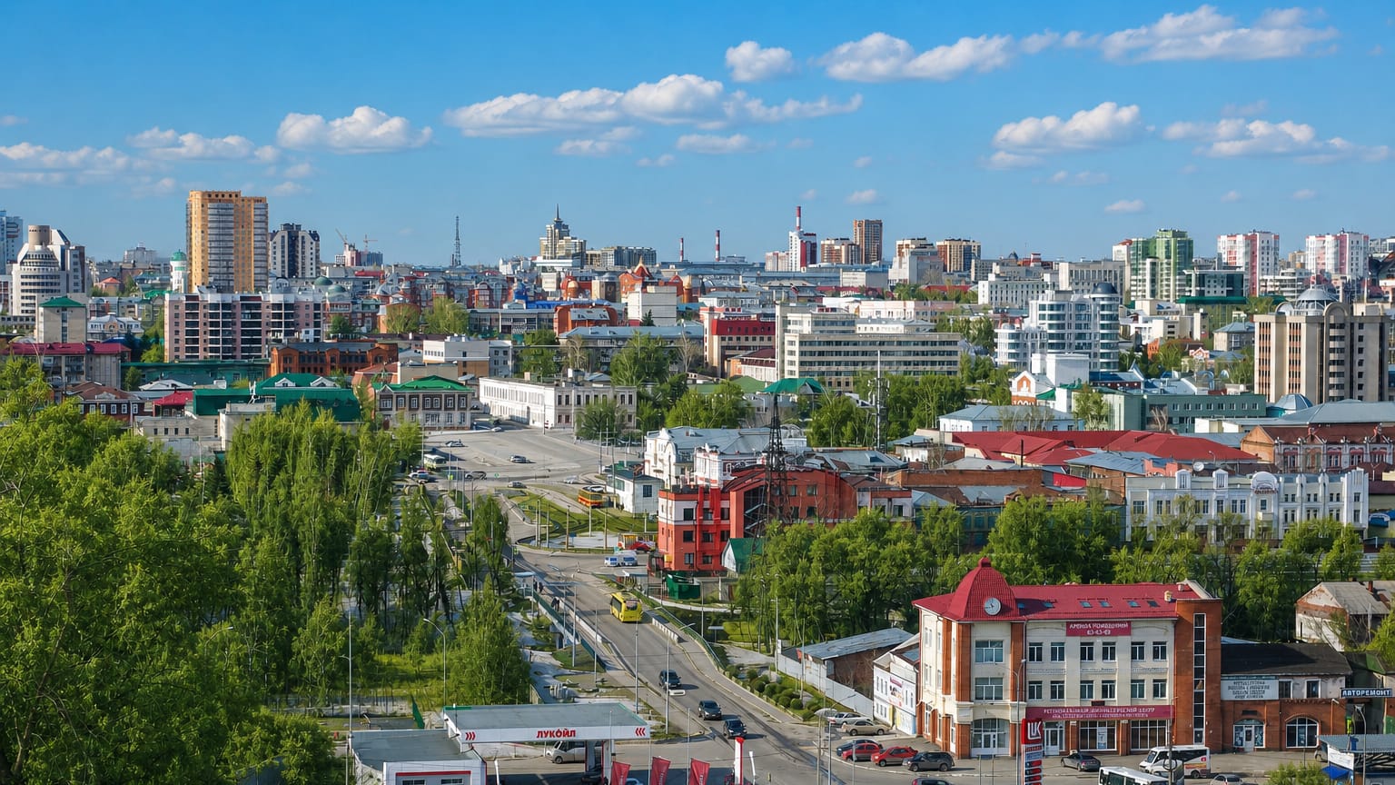 Wide daytime panorama of Barnaul, Russia, with historic rooftops and apartment towers beyond leafy streets.