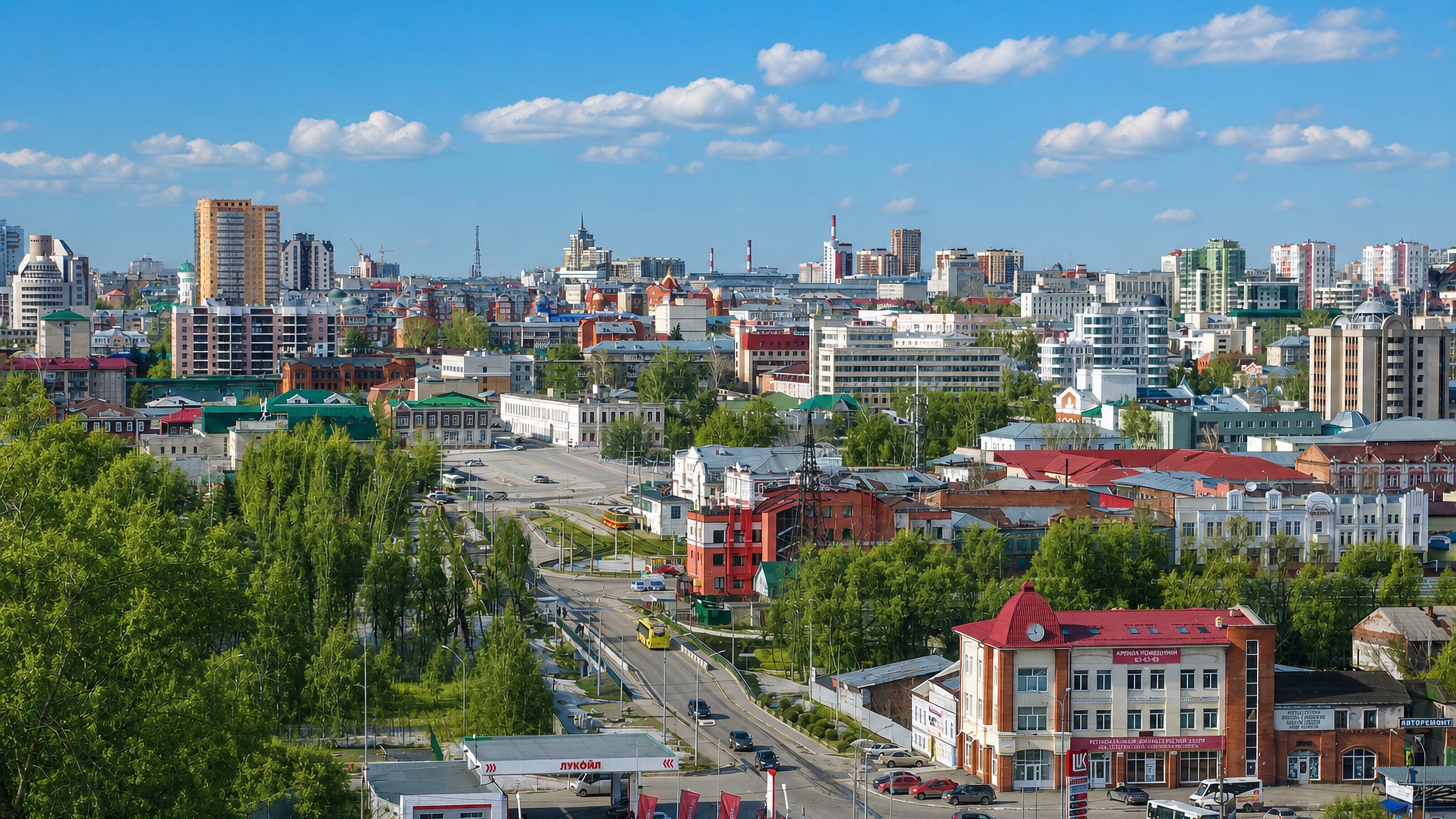 Wide daytime panorama of Barnaul, Russia, with historic rooftops and apartment towers beyond leafy streets.