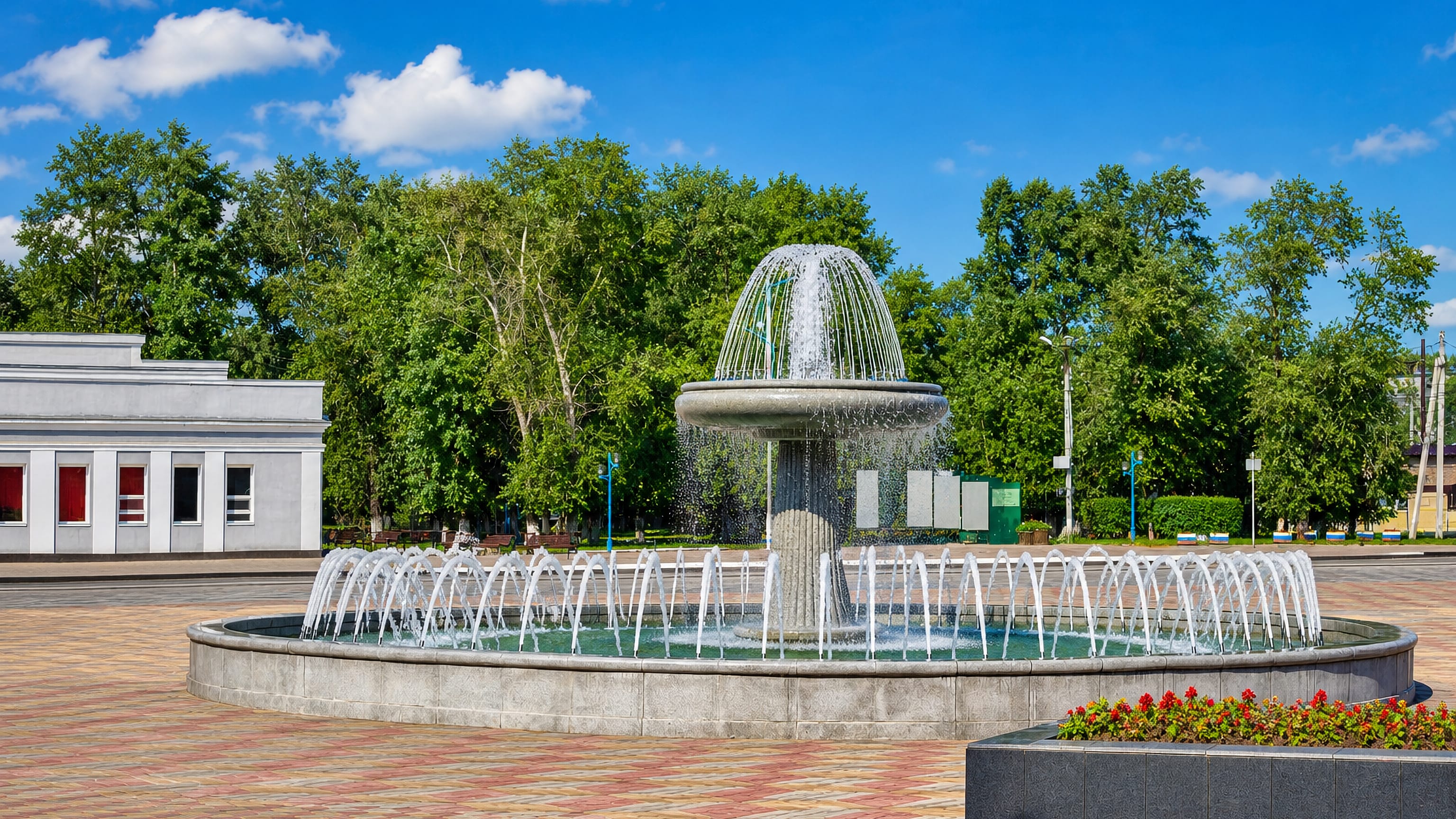 Tiered fountain on a paved town square in Belogorsk, Russia, framed by summer birches.