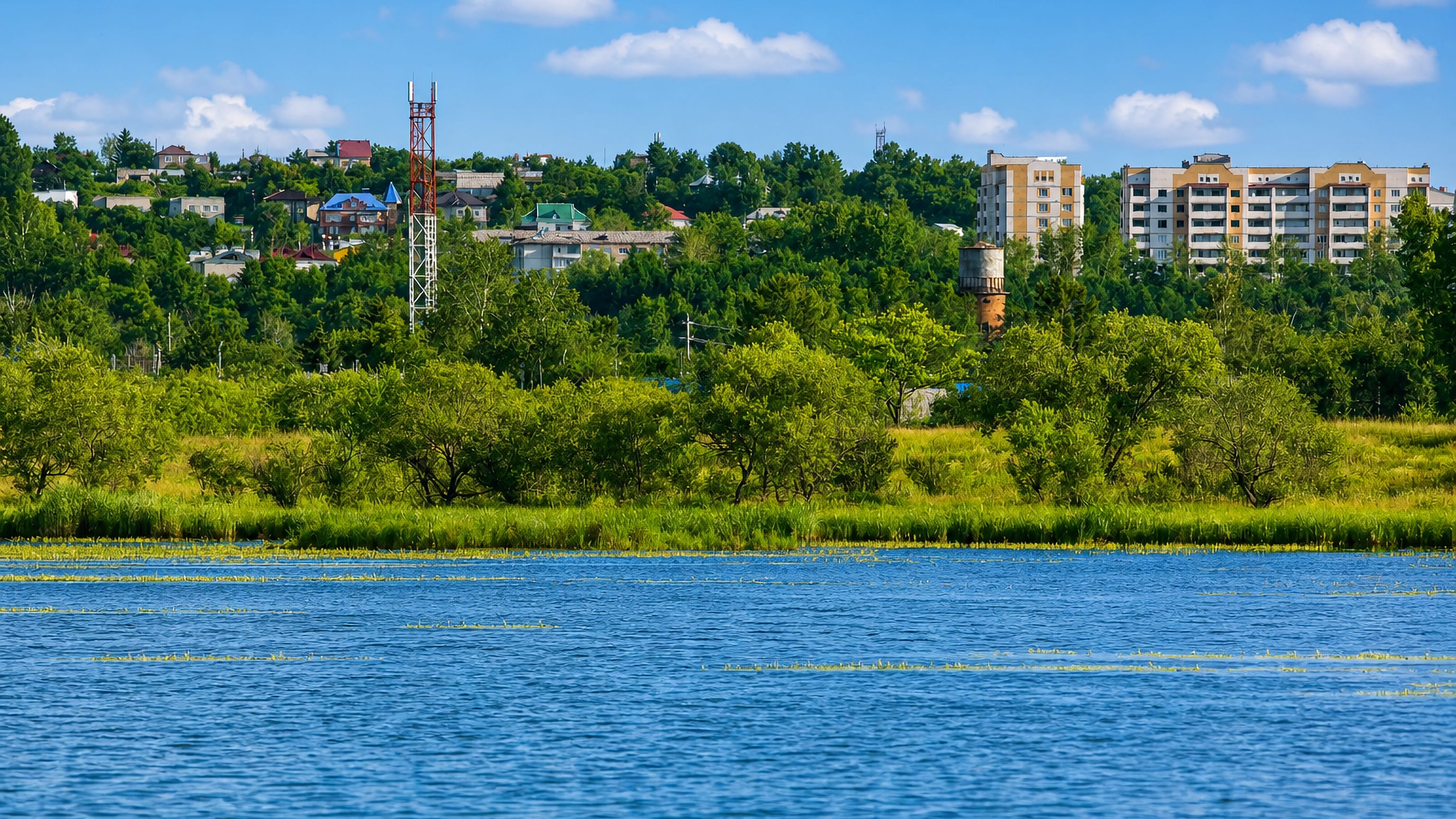 Apartment blocks and houses of Svobodnyy on a wooded riverbank above calm blue water in Amur Oblast.