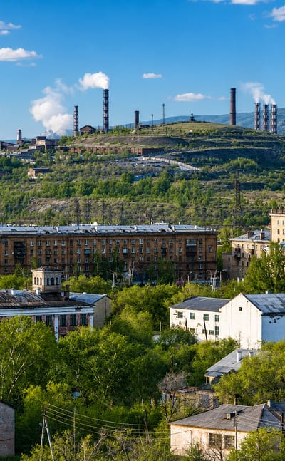 Magnitogorsk townscape with mid-rise apartments below the hillside smokestacks of its steelworks under a blue sky.