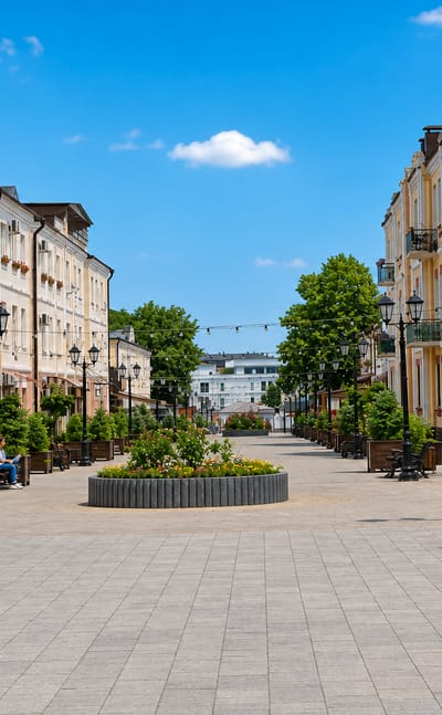 Pedestrian promenade in central Nalchik lined with pastel low-rise buildings, planters and street lamps under a blue sky.