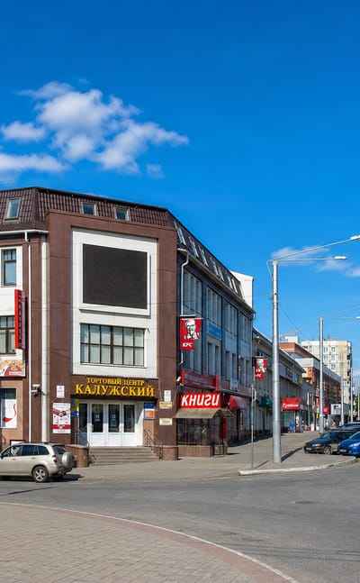Central Kaluga street corner with a brick commercial building, shops and passing traffic under a blue sky.