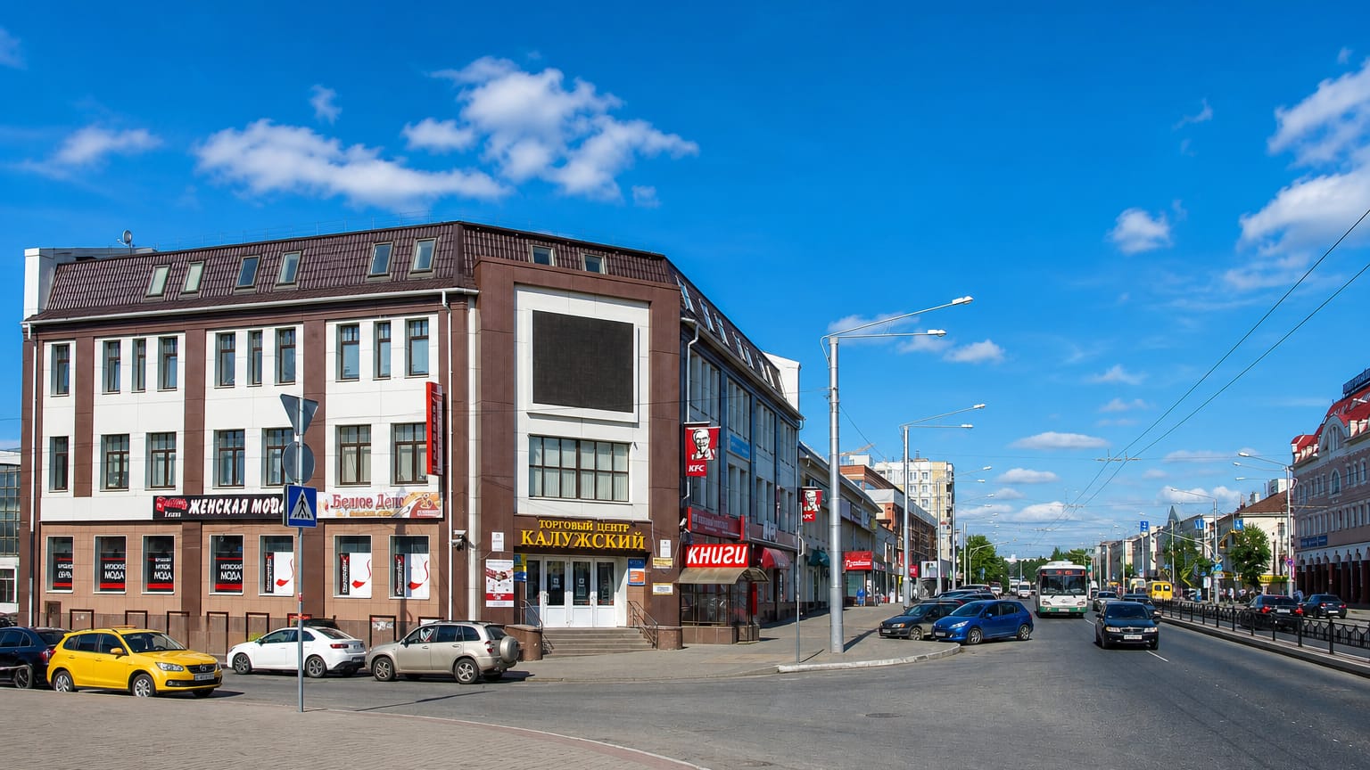 Central Kaluga street corner with a brick commercial building, shops and passing traffic under a blue sky.