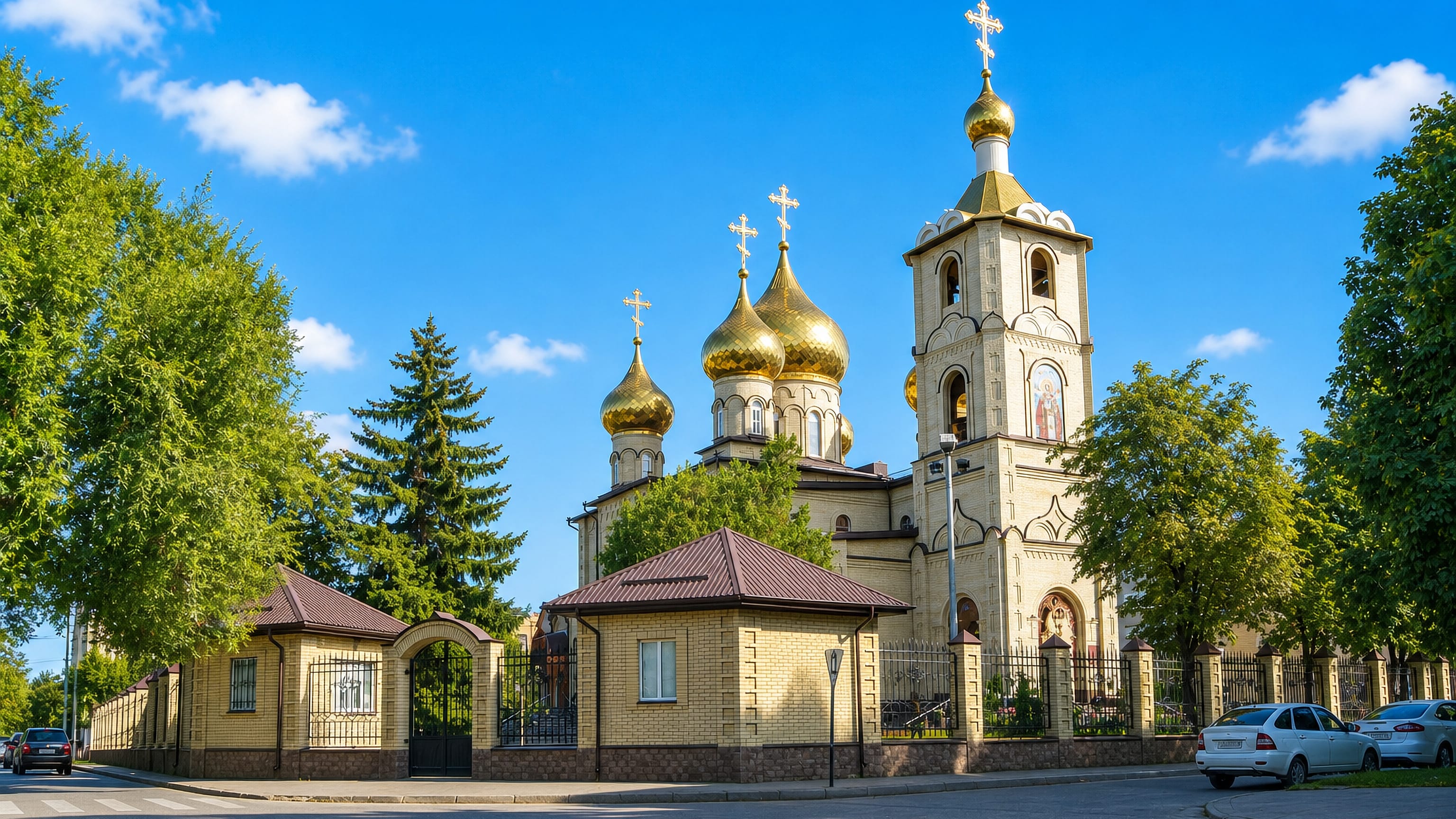 A golden-domed Orthodox church and bell tower behind a brick fence on a sunlit street corner in Cherkessk