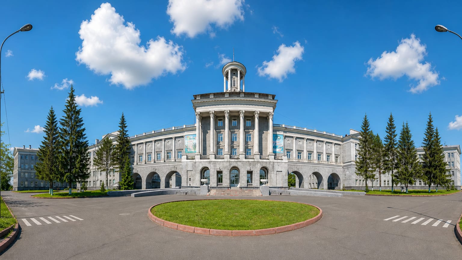 Grand columned Stalinist university building framed by fir trees and a circular lawn in Kemerovo, Russia.