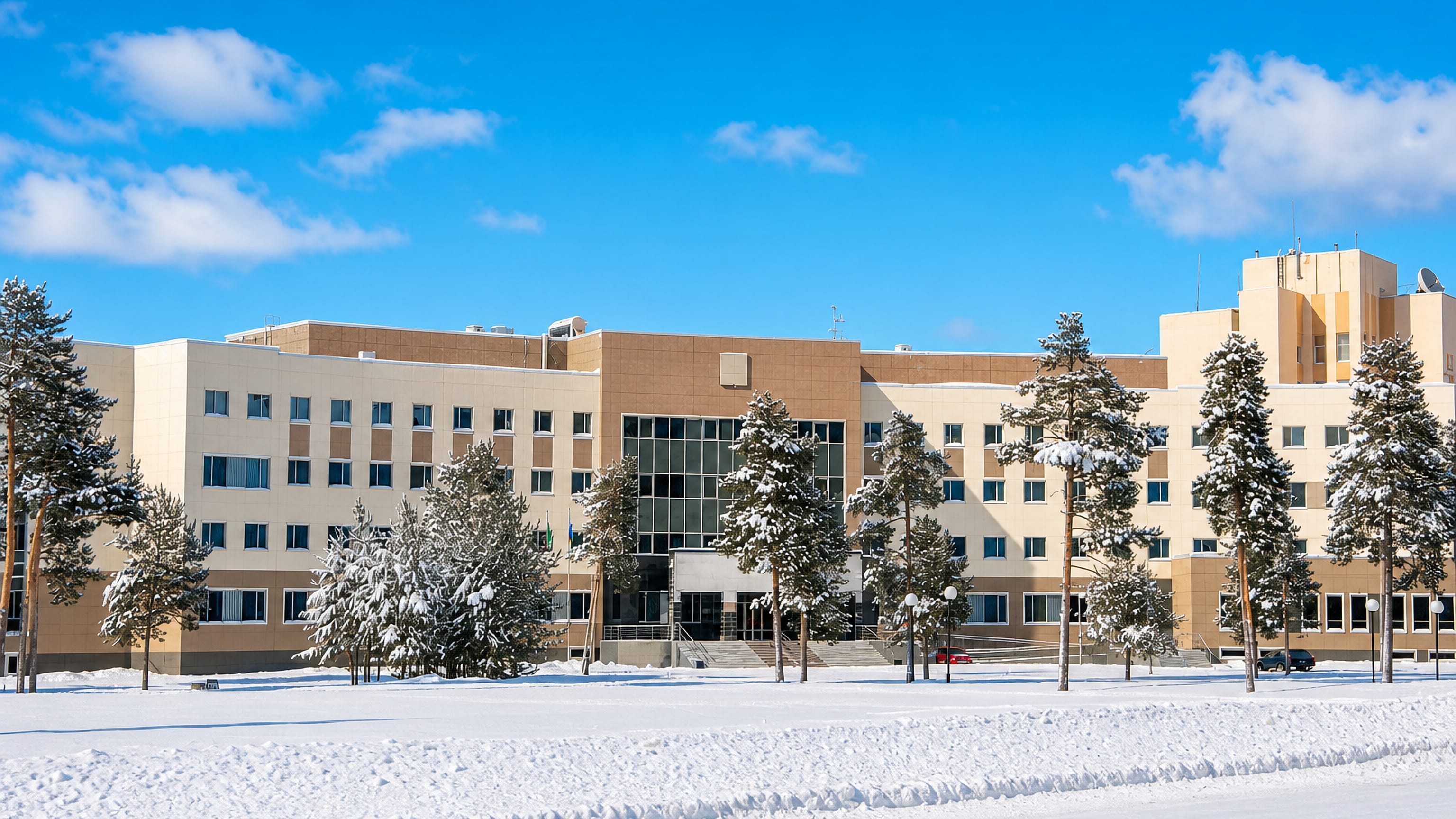 A modern civic building in Kogalym behind snow-covered pines and deep winter snow under clear skies.