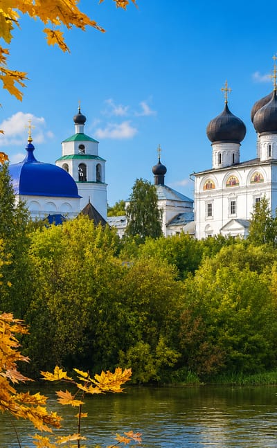 Kirov's white Trifonov Monastery with black onion domes across the river, framed by golden autumn foliage.