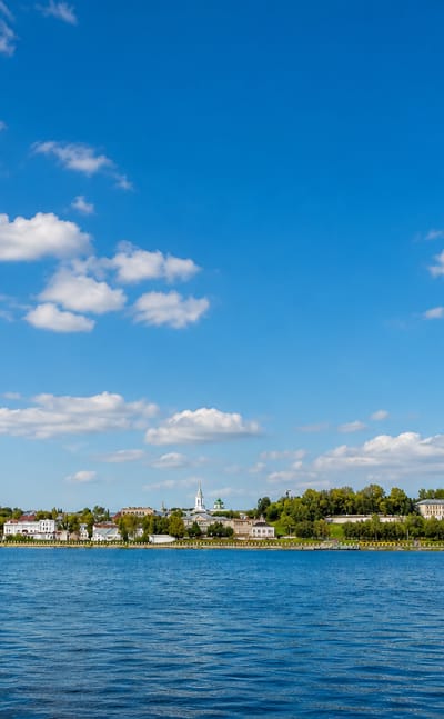 Kostroma's tree-lined Volga River waterfront seen from the water under a wide cloud-dotted sky.