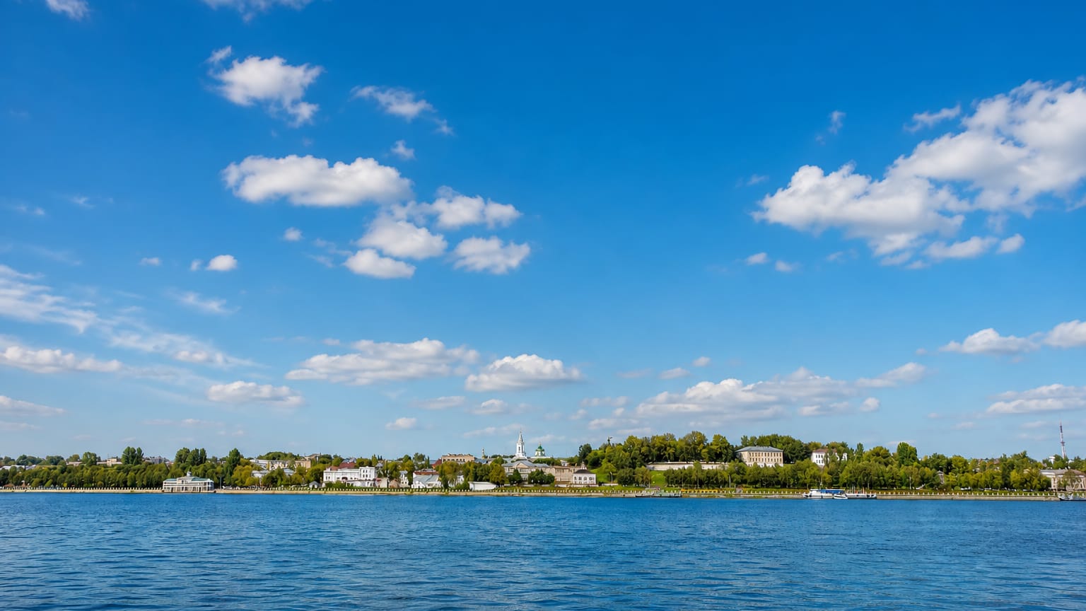 Kostroma's tree-lined Volga River waterfront seen from the water under a wide cloud-dotted sky.
