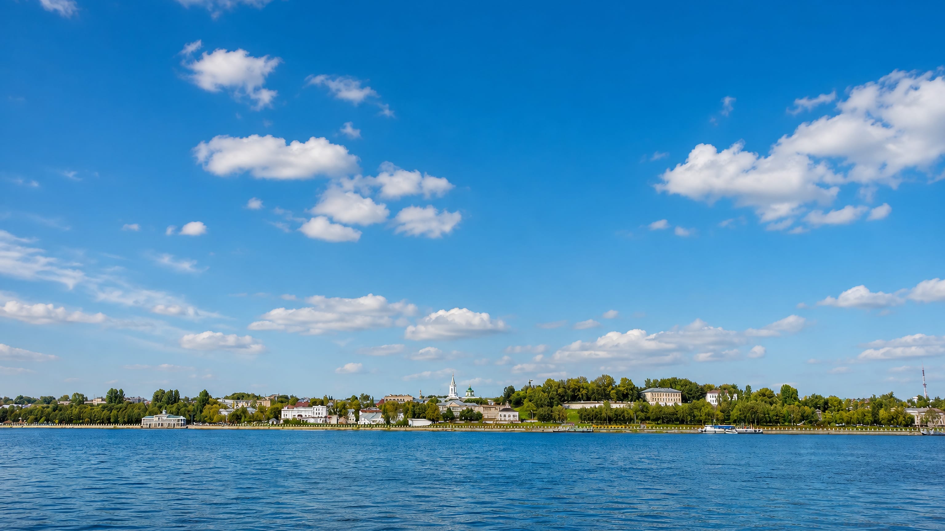 Kostroma's tree-lined Volga River waterfront seen from the water under a wide cloud-dotted sky.