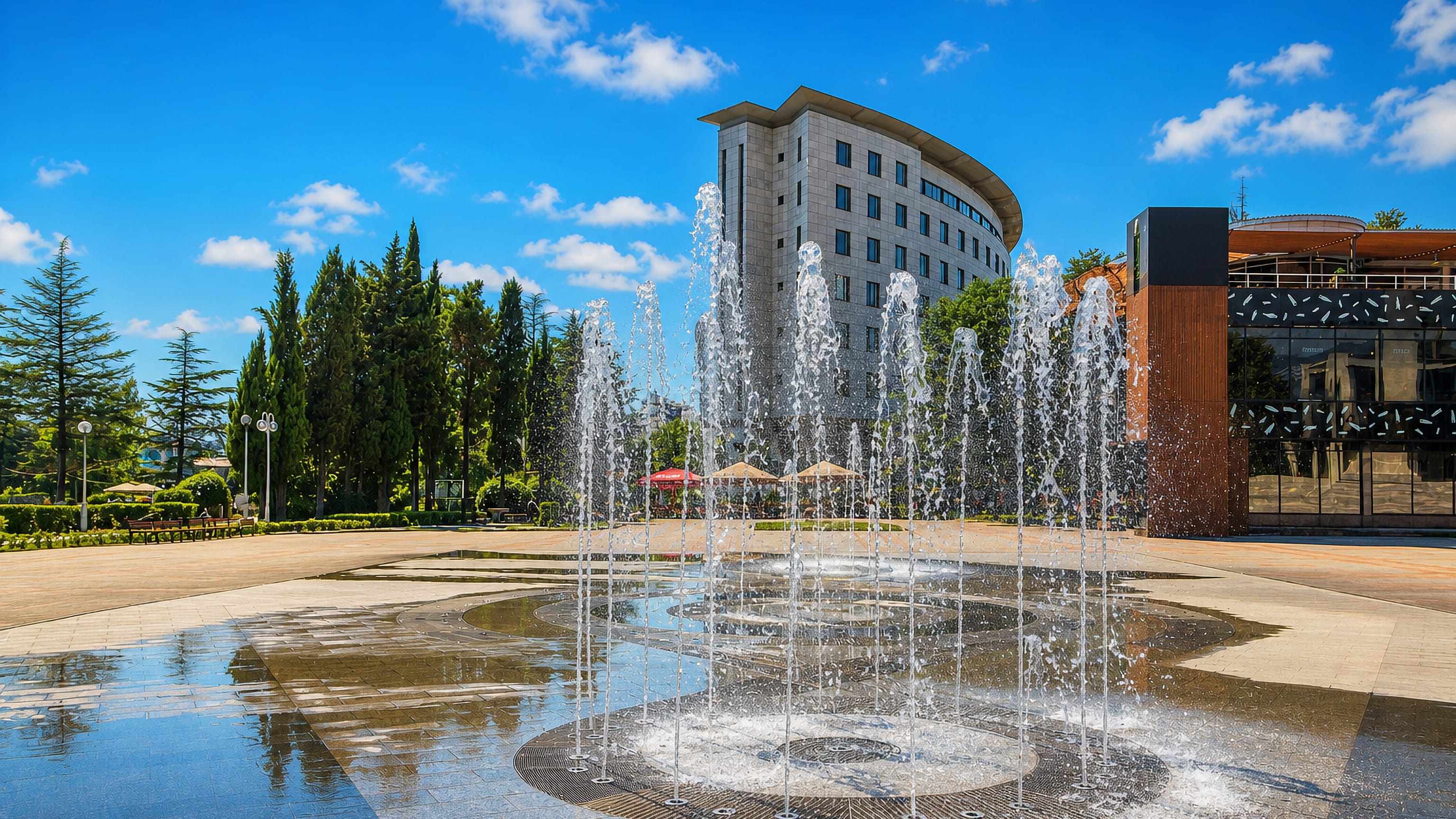A paved plaza fountain in Tuapse, Russia, backed by a curved modern hotel building and tall cypress trees.
