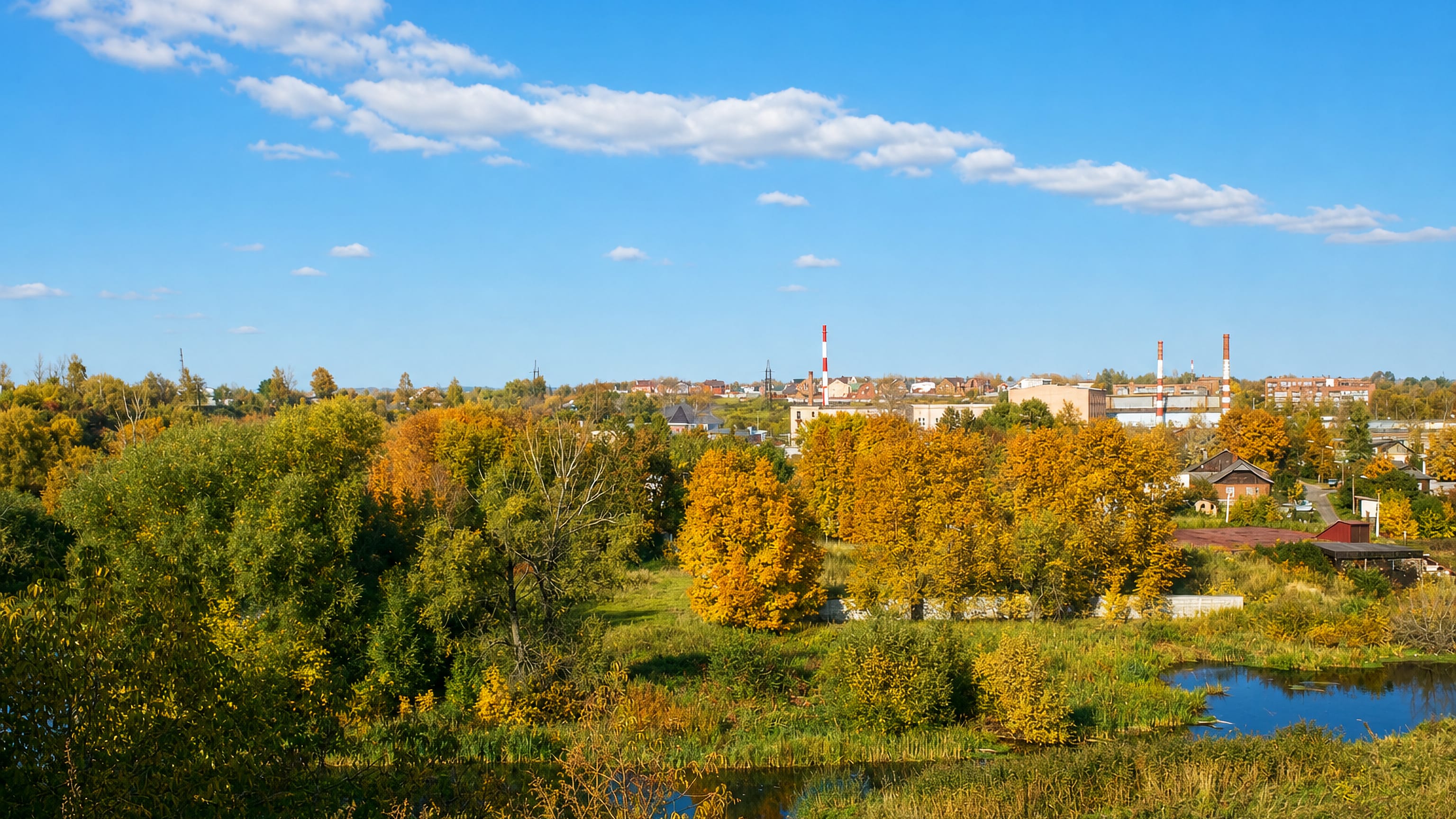 Autumn trees and a pond in front of the low skyline and factory chimneys of Klin, Moscow Oblast.