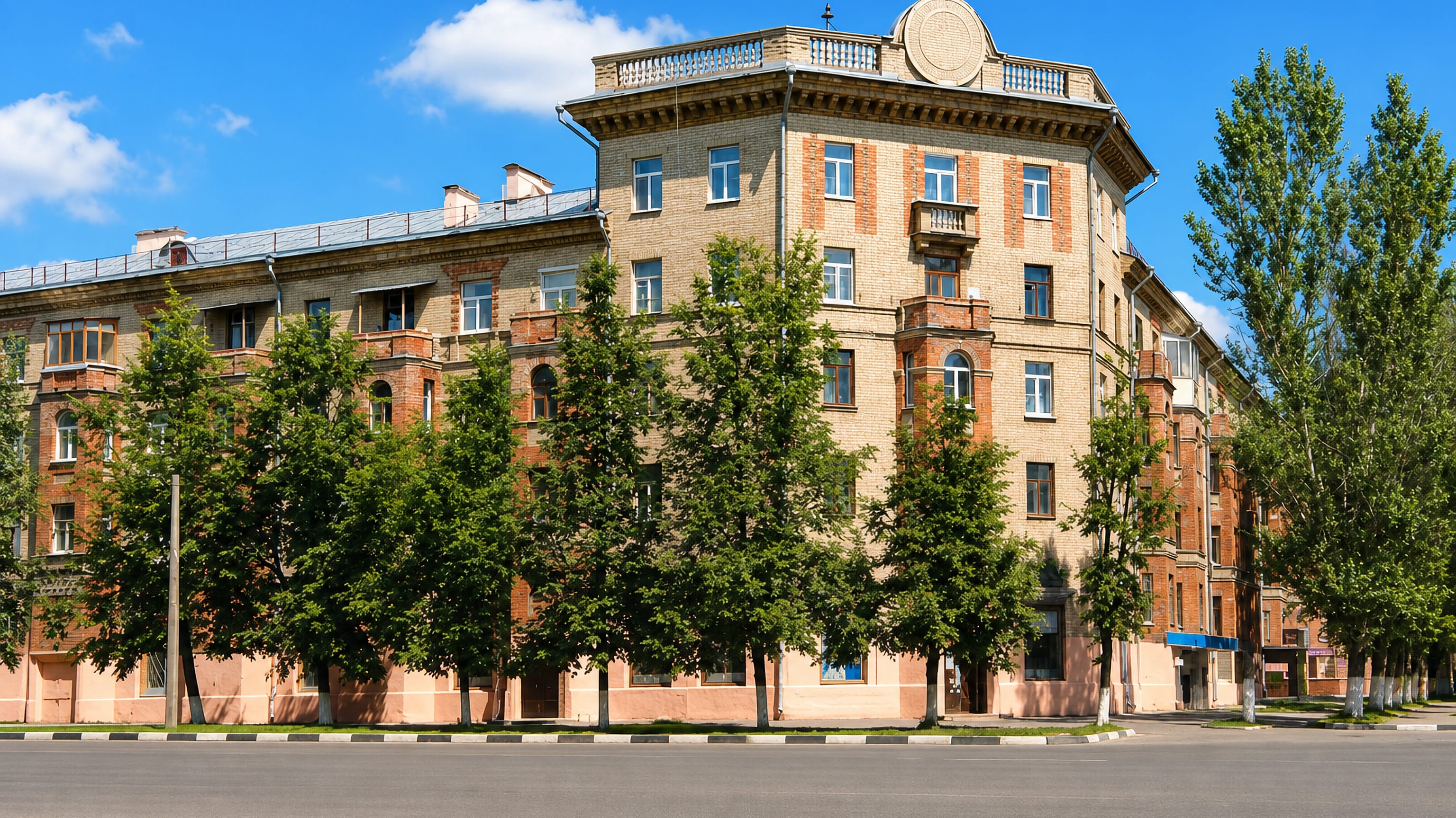 A Stalin-era brick corner apartment block behind leafy trees on a wide street in Lytkarino, Russia.