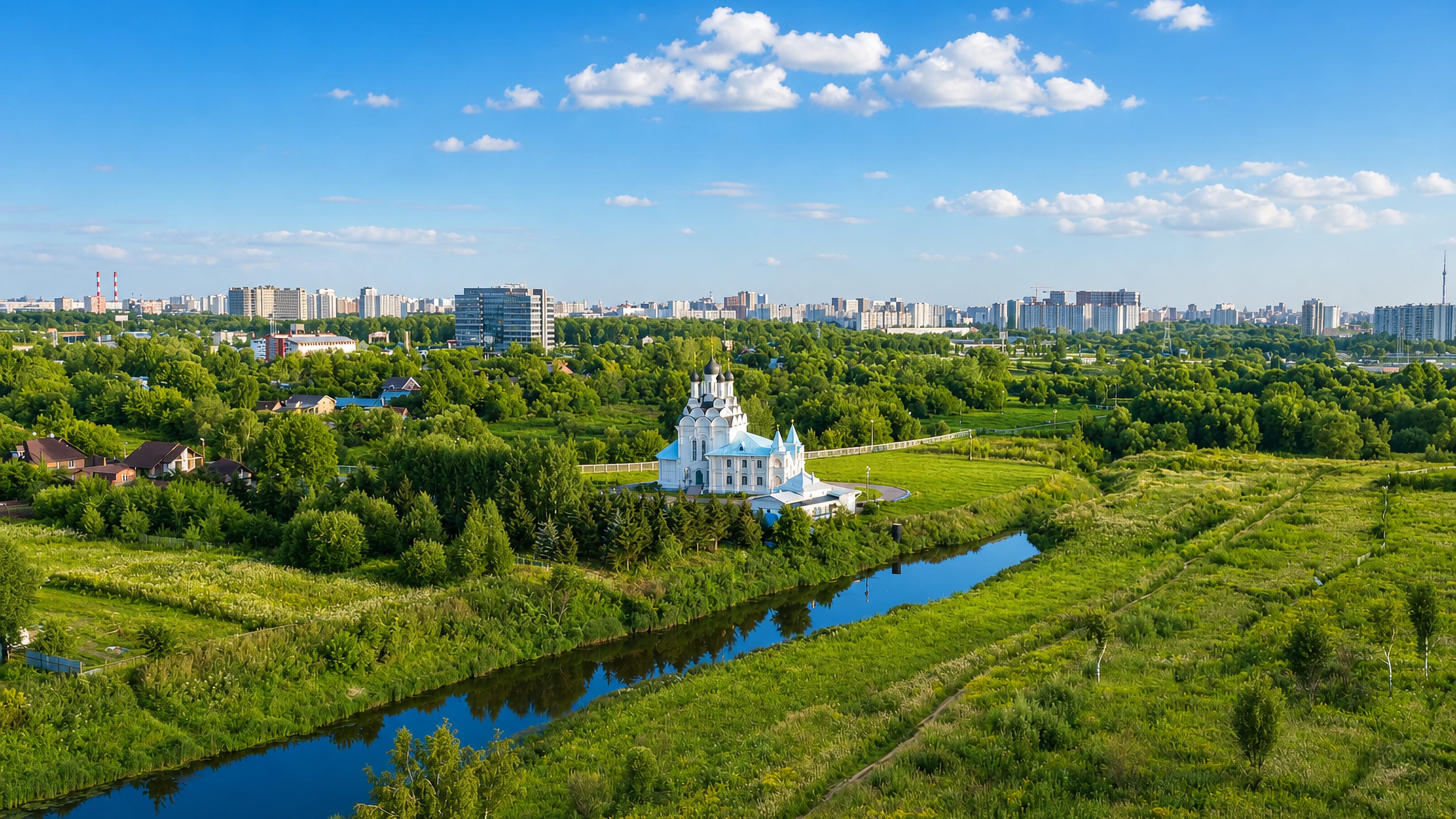 White Orthodox church on a green river meadow below the apartment-block skyline of Mytishchi, Russia.