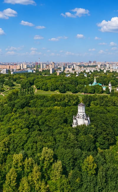 Aerial of Kolomenskoye park in Moscow with the white Church of the Ascension among trees and the city skyline beyond.