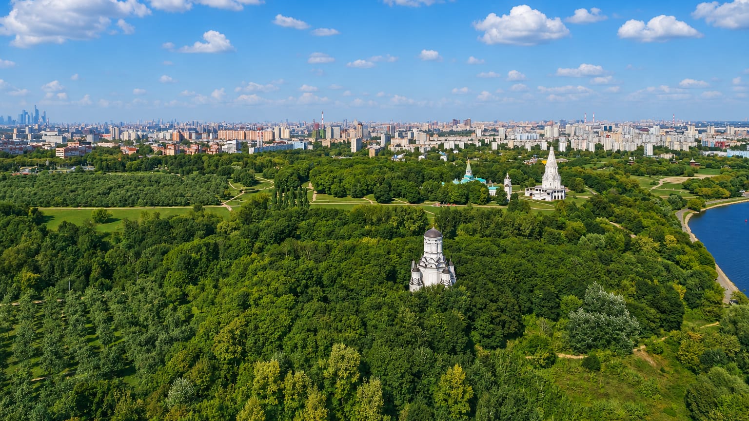 Aerial of Kolomenskoye park in Moscow with the white Church of the Ascension among trees and the city skyline beyond.