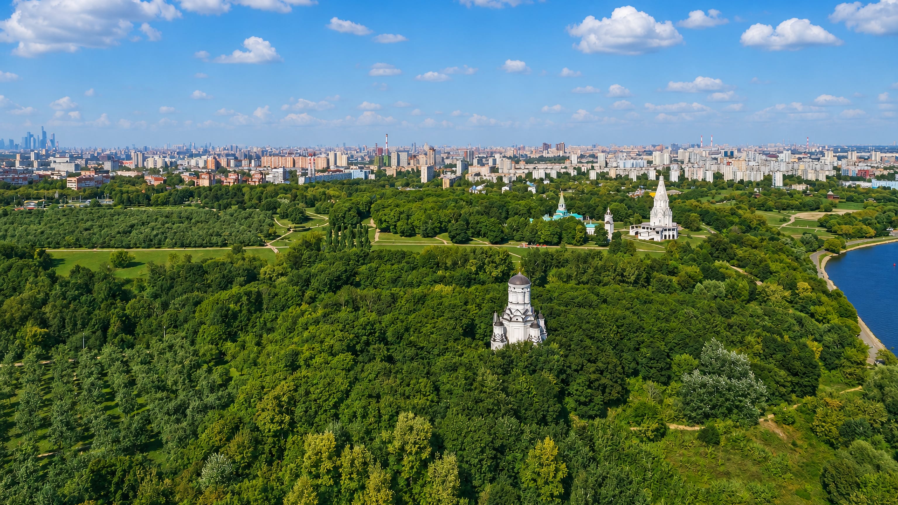 Aerial of Kolomenskoye park in Moscow with the white Church of the Ascension among trees and the city skyline beyond.