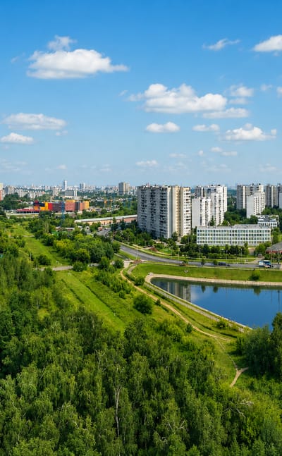 Green outer Moscow district Vykhino-Zhulebino with a pond, wooded parkland and residential tower blocks under a blue sky.