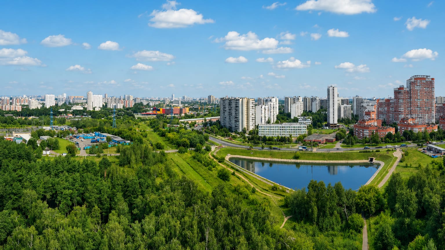 Green outer Moscow district Vykhino-Zhulebino with a pond, wooded parkland and residential tower blocks under a blue sky.