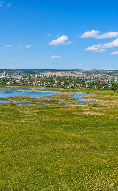 Iskitim, Russia, seen across a grassy meadow and pond, with apartment blocks and cement works chimneys beyond.