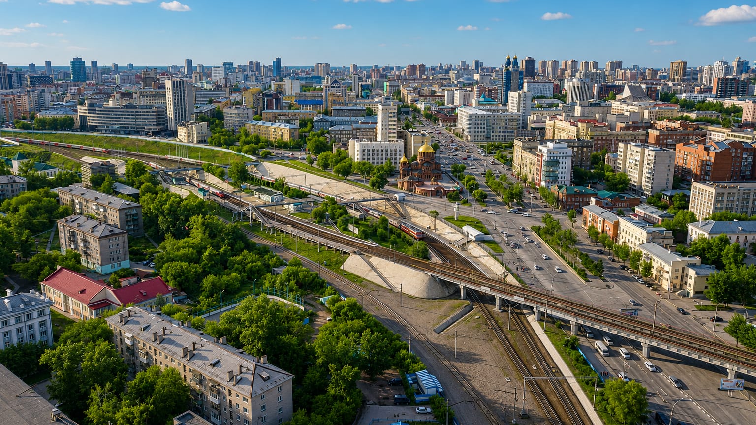 Aerial view of central Novosibirsk with rail lines, a road interchange, a golden-domed church and a dense mid-rise skyline.