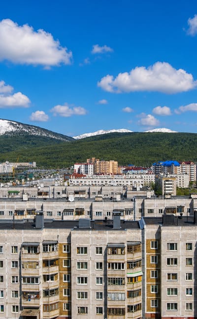 Apartment blocks of Yuzhno-Sakhalinsk, Russia, below forested, snow-streaked mountains under a blue sky.
