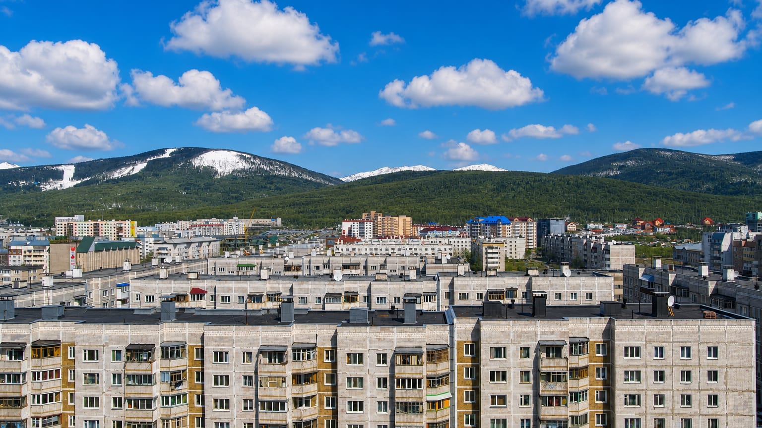 Apartment blocks of Yuzhno-Sakhalinsk, Russia, below forested, snow-streaked mountains under a blue sky.