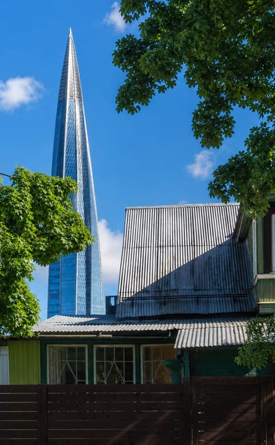 The Lakhta Center skyscraper towering behind an old wooden dacha and green trees in Lakhta, Saint Petersburg.
